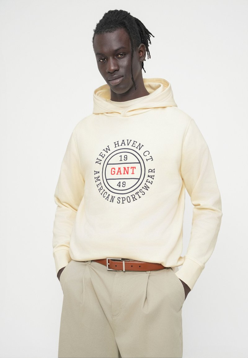 Sweat à capuche crème avec logo avant, affichant "GANT" en rouge dans un design circulaire. Associé à un pantalon beige et une ceinture marron.