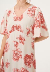 Femme portant une robe blanche à imprimé floral rouge, col en V et manches courtes volantées, visible des épaules jusqu'à mi-cuisse.