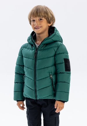 Veste rembourrée verte avec capuche, munie d'une fermeture éclair, de poches latérales et d'un patch texturé noir sur la manche gauche.