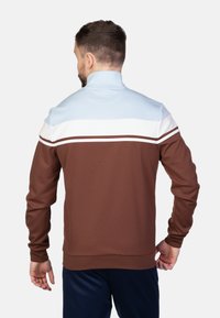 Bruine zip-up jas met lichtblauwe en witte horizontale strepen over de schouders. Beschikt over een hoge kraag en rekbare stof.