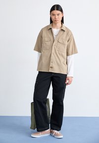 Beige short-sleeve shirt with two chest pockets, black cargo pants with side zipper pockets, and light pink sneakers. Olive green tote bag.