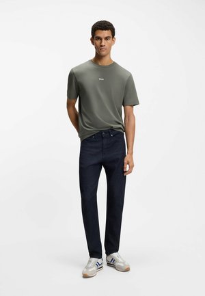 Dunkle Jeans aus Denim kombiniert mit einem figurbetonten olivgrünen T-Shirt mit weißem Logo-Schriftzug. Weiße und graue Sneaker vervollständigen das Outfit.