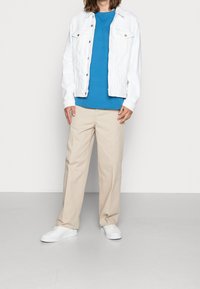Giacca in denim bianco sopra una t-shirt blu, abbinata a pantaloni beige e sneakers bianche. Tessuto morbido, taglio rilassato, con dettagli a bottoni.