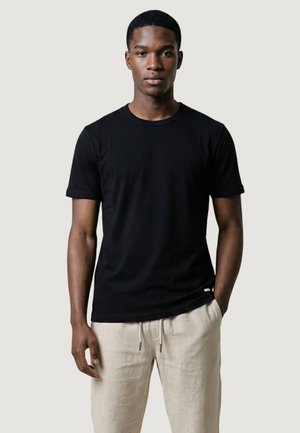 Giovane uomo che indossa una maglietta nera a maniche corte semplice e pantaloni beige chiaro con coulisse, in piedi contro uno sfondo chiaro uniforme.