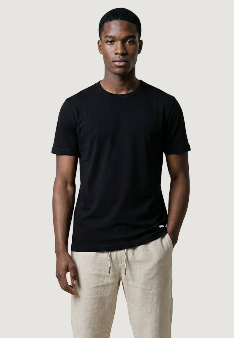 Giovane uomo che indossa una maglietta nera a maniche corte semplice e pantaloni beige chiaro con coulisse, in piedi contro uno sfondo chiaro uniforme.