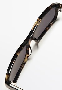 Sluneční brýle s motivem želvoviny a tmavými skly. Rám má hladké hrany a elegantní, zaoblený design s kovovými akcenty poblíž pantů.