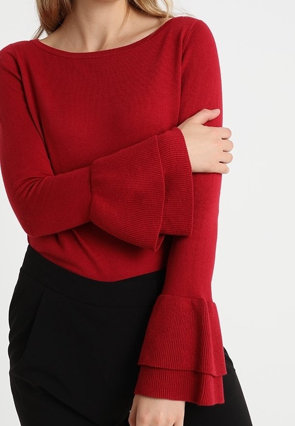 Femme portant un pull rouge à manches longues avec des manches cloches superposées et un pantalon noir taille haute sur un fond uni.