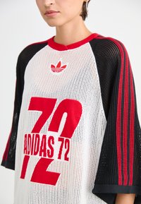 Persona, kas valkā baltu Adidas 72 jersiju ar melniem piedurknēm, uz kurām ir sarkanas svītras un Adidas logo tuvu kakla izgriezumam.