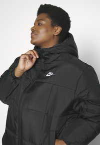 Doudoune noire avec capuche, dotée d'une fermeture éclair à l'avant et de poignets élastiques. Logo Nike blanc notable sur la poitrine gauche.