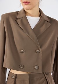 Krótkie brązowe marynarka z gładkiej tkaniny, z podwójnym zapięciem na guziki i czterema okrągłymi guzikami. Charakterystyczne szerokie klapy i dopasowany krój.