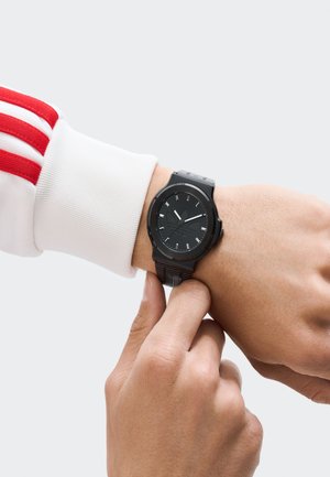 Cilvēks, kurš valkā melnu analogās pulksteņa ar Adidas logo, pielāgo pulksteni uz savas kreisās rokas, ģērbies baltā piedurknē ar sarkaniem svītrām.