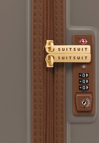 Bruine kofferrits met dubbele gouden trekker met het label "SUITSUIT," voorzien van een combinatie-slot en een rode TSA-sleutelgaatje-indicator.