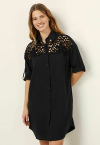 Rochie neagră cu butoni în față, mâneci scurte rulate și un overlay din dantelă croșetată pe umeri și piept.