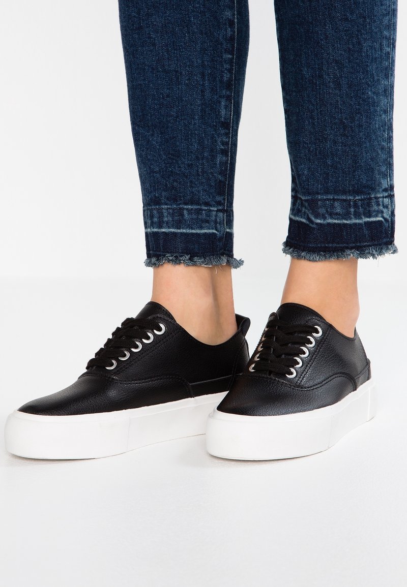 Baskets en cuir noir avec un bout arrondi, un laçage à l'avant et une semelle plateform blanche. Portées avec un jean frangé bleu foncé.