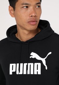 Hoodie noir en mélange de coton ; présente le logo « PUMA » blanc et un graphique de puma en train de sauter. Comprend des cordons de serrage et une poche kangourou.