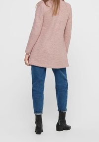 Cappotto rosa texturizzato con maniche lunghe, orlo leggermente svasato, indossato su jeans blu e stivali neri alla caviglia, visto da dietro.
