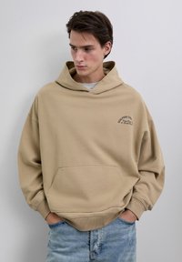 Jonge man met donker haar kijkt omlaag terwijl hij een beige oversized hoodie met een voorzak en blauwe jeans draagt, tegen een eenvoudige lichte muur.