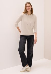 Kvinna som ler, klädd i en ljusbeige hoodietopp med 3/4-långa ärmar, svarta vida jeans och vita sneakers med svarta detaljer.