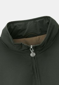 Veste vert foncé avec un col haut et une fermeture éclair argentée. L'intérieur est beige, avec une texture lisse et un design minimaliste.