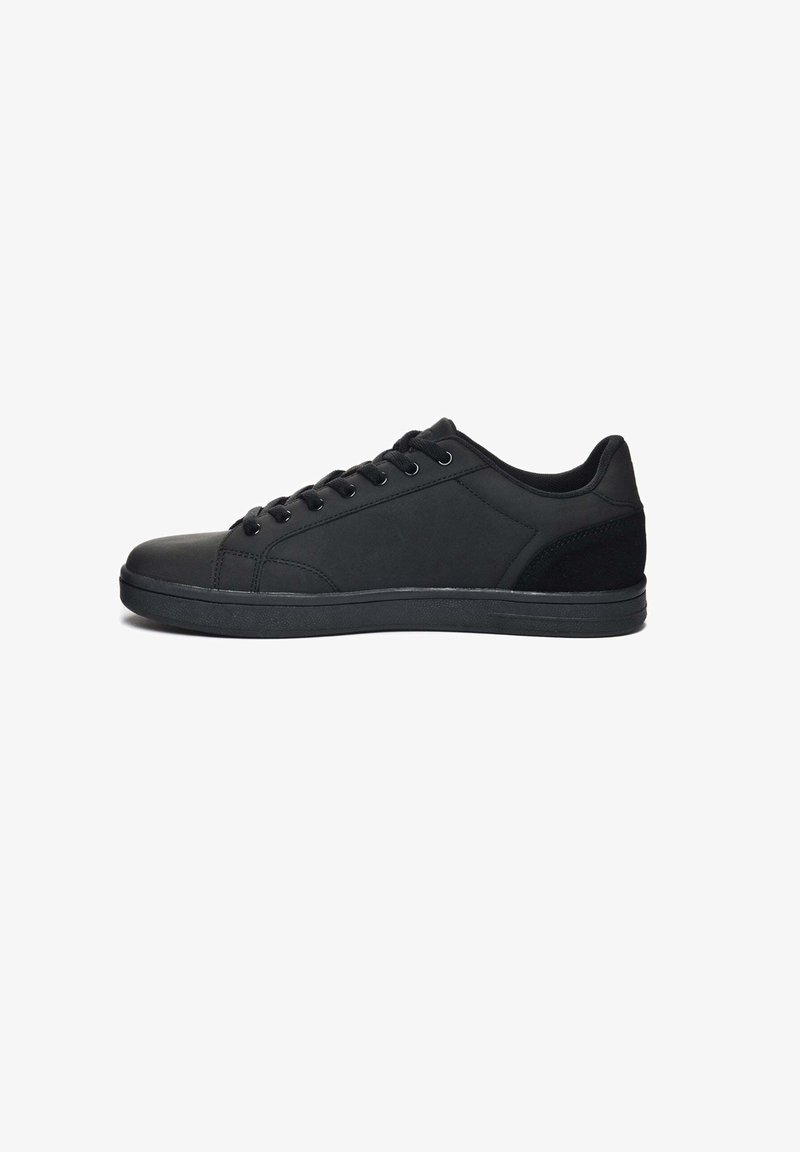 Zapatilla deportiva negra con parte superior de cuero suave, puntera redonda, suela plana, seis ojales y un acento en el talón texturizado. Diseño simple y de corte bajo.