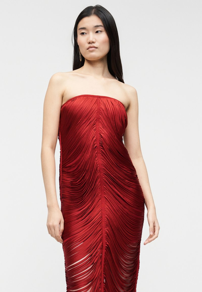 Jeune femme aux longs cheveux noirs portant une robe rouge sans bretelles, froncée avec une texture plissée verticale, sur un fond gris clair.