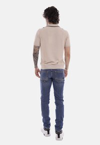Homme aux cheveux bouclés et aux bras tatoués, portant une chemise beige à manches courtes, un jean bleu et des baskets blanches, debout de dos.