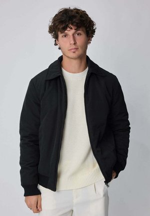 Jeune homme aux cheveux bouclés portant une veste noire sur un pull blanc et un pantalon blanc, debout devant un fond clair uni.