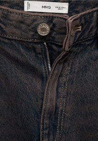 Jean en denim bleu foncé avec une fermeture éclair et un bouton métallique, présentant une texture subtile et des détails de couture, étiquette de la marque visible à l'intérieur.