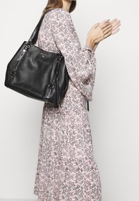 Sort tekstureret læder totebag med to skulderstropper, metalhardware og frynse-detalje, båret sammen med en mønstret pink kjole.