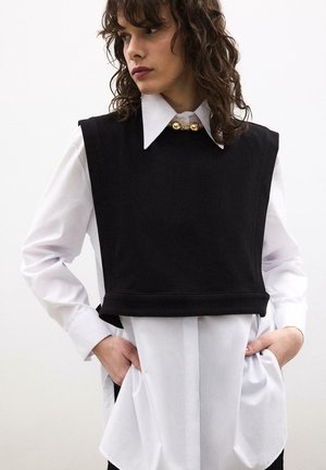 Žena s kučeravými vlasmi má na sebe bielu košeľu s veľkým golierom a čiernu bezrukávovú vestu, ruky v kapse, pozerá doľava na bielom pozadí.