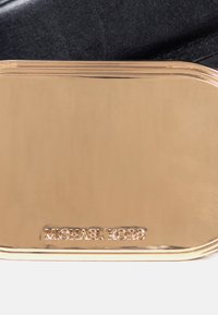 Plaque métallique dorée rectangulaire brillante avec des bords arrondis gravée "MICHAEL KORS" sur un fond en tissu noir.