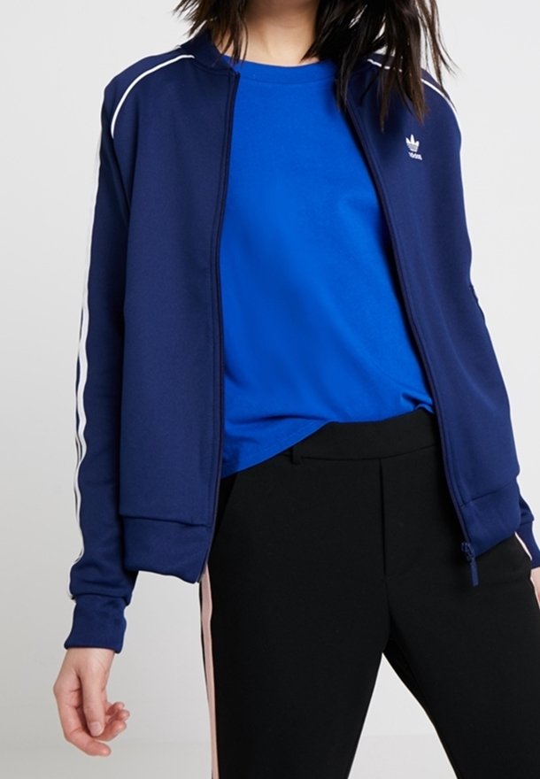 Marineblauwe sportjas met witte strepen langs de mouwen, ritssluiting, geribbelde manchetten en zoom, gecombineerd met een blauw shirt en zwarte broek.