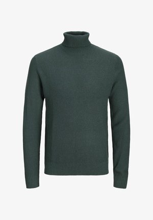 Persona che indossa un maglione a collo alto a nido d'ape verde scuro, jeans blu scuro e sneakers bianche, in piedi contro uno sfondo semplice.