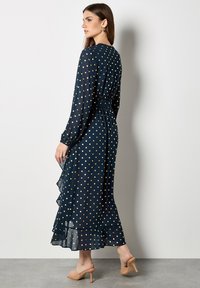 Lungo abito blu navy con maniche lunghe, motivo a pois dorati, vita arricciata, orlo arricciato e texture trasparente, abbinato a tacchi nudi.