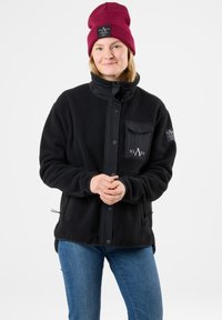 Giacca in pile nera con colletto alto, bottoni a pressione e taschino sul petto. Abbinata a jeans blu e un berretto bordeaux. Logo del brand in evidenza.