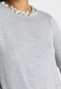 Pull gris avec un col rond orné de grappes de perles blanches et argentées. Présente une texture douce et une coupe classique.