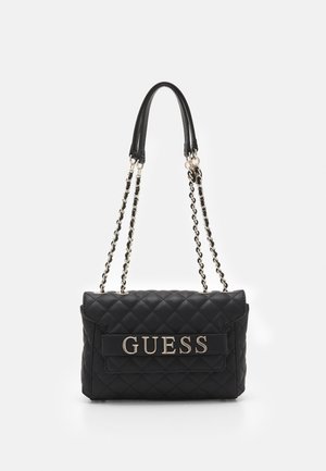 Sac à main noir matelassé avec double bandoulière chaîne, présentant un logo métallique "GUESS" proéminent sur un rabat avant. Texture lisse et forme compacte.