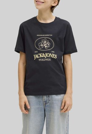 Persona vistiendo una camiseta negra de Jack & Jones con un diseño floral y texto beige, combinada con jeans de mezclilla azul claro sobre un fondo gris.