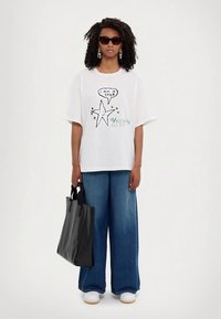 Moteris, dėvinti akinius nuo saulės, baltą per didelį marškinėlį su žvaigždės grafika ir tekstu, plačių kojų džinsus, baltus sportbačius, laiko juodą rankinę.