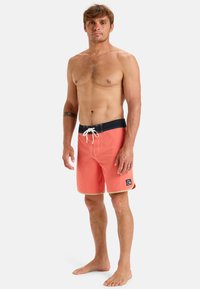 Korálové plavky s námořnickým pasem a bílou šnůrkou. Obsahují postranní nášivku s logem a béžový lem na spodním okraji. Hladká struktura materiálu.