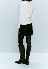 Czarne szorty, biała prążkowana bluzka z długim rękawem, teksturowane czarne kozaki do kolan na masywnej podeszwie, modelka zwrócona tyłem na jasnym tle.