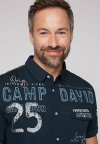 Short-sleeve dark blue button-up shirt featuring various embroidered text, including "CAMP DAVID" and "25," with a chest pocket detail.