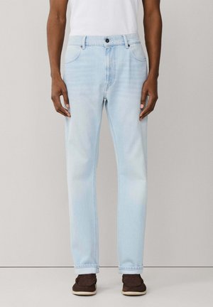 Homme portant un jean droit bleu clair, une chemise blanche et des chaussures marron foncé, debout devant un fond clair uni.