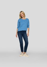 Eine Frau steht und trägt ein strukturiertes, blaues Langarmoberteil, dunkle Jeans und cremefarbene Sneakers vor einem schlichten, hellgrauen Hintergrund.