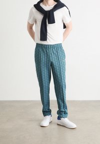 Pánské vzorované tepláky v zelené barvě s geometrickým vzorem, elastickým pasem a modrými bočními pruhy. Nošeny s bílým tričkem a námořnicky modrým svetrem.