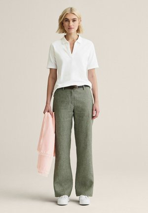 Kvinde i hvid kortærmet polo, grønne vidde bukser, hvide sneakers, holder et lyserødt tørklæde, stående på en beige baggrund.