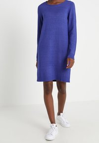 Una donna indossa un vestito maglione blu, lungo fino al ginocchio e con maniche lunghe, abbinato a sneakers bianche, in piedi su uno sfondo bianco uniforme.