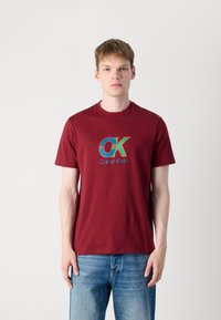 Bordó pamut póló kerek nyakkivágással; a frontrészen egy színes "OK" logó és a "Calvin Klein" felirat található. Világos farmernadrággal viselve.