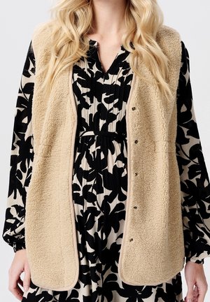 Beżowa kamizelka z polaru sherpa na czarno-białej sukience w kwiaty z długimi rękawami, taliowanym stanem i dekoltem w kształcie litery V.