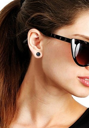Boucles d'oreilles noires en forme circulaire, fabriquées dans un matériau lisse, portées par un modèle avec des lunettes de soleil noires réfléchissantes et des cheveux bruns lisses.
